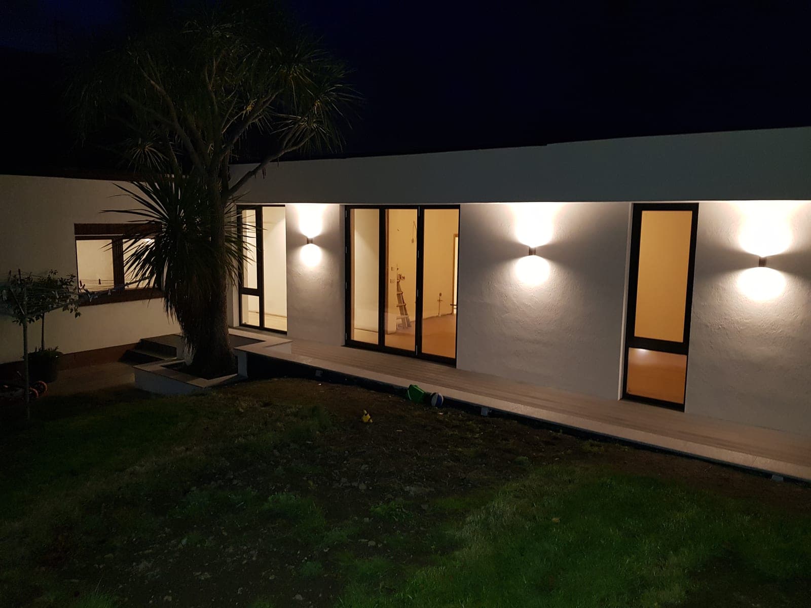 Single-storey home lit at night, with wall lights along the white render